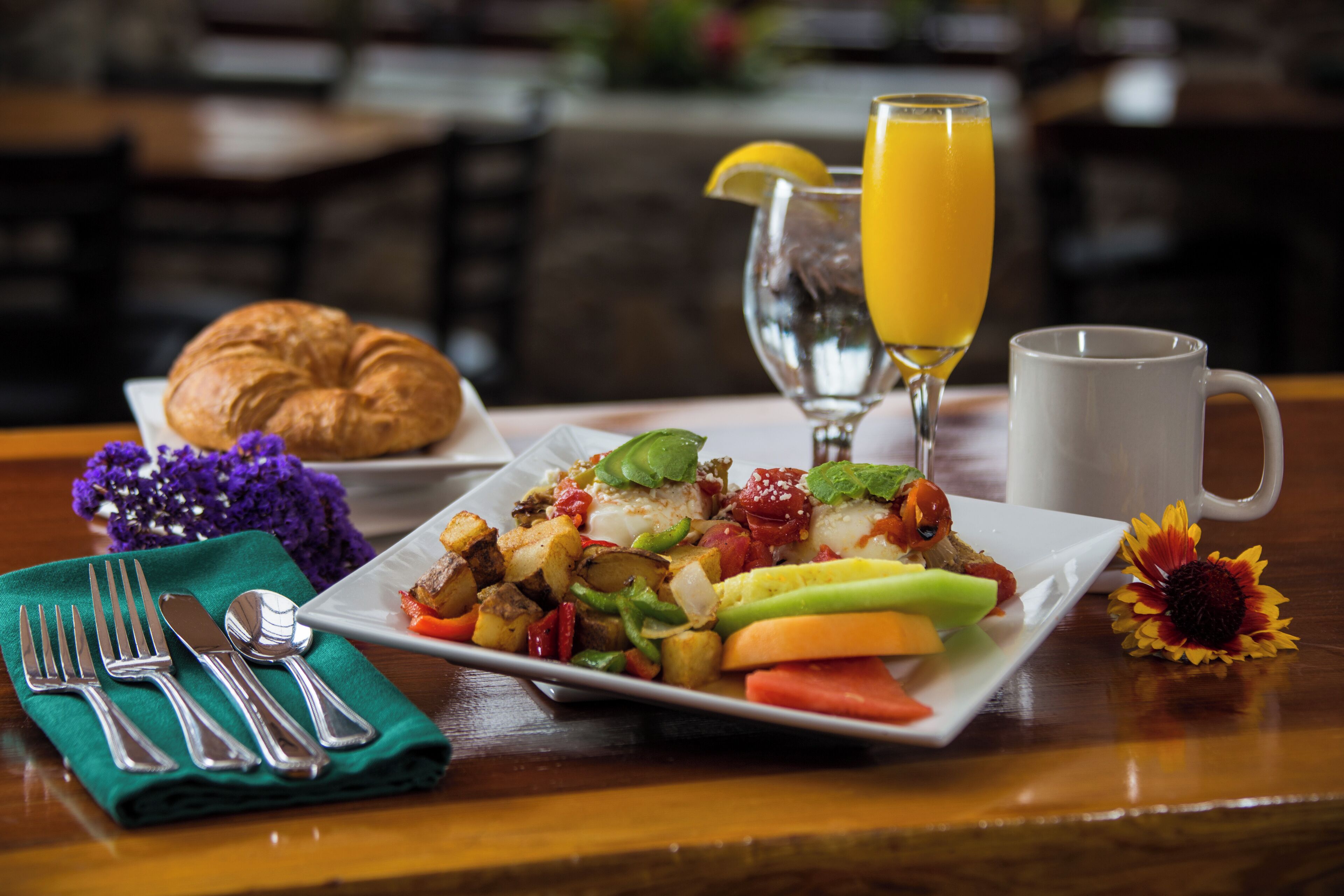 A breakfast setting with a croissant, a plate of vegetables, a glass of orange juice, and a mug on a wooden table.