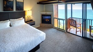 A hotel room with a large bed, a fireplace, a balcony with a view of the ocean, and two framed pictures on the wall.