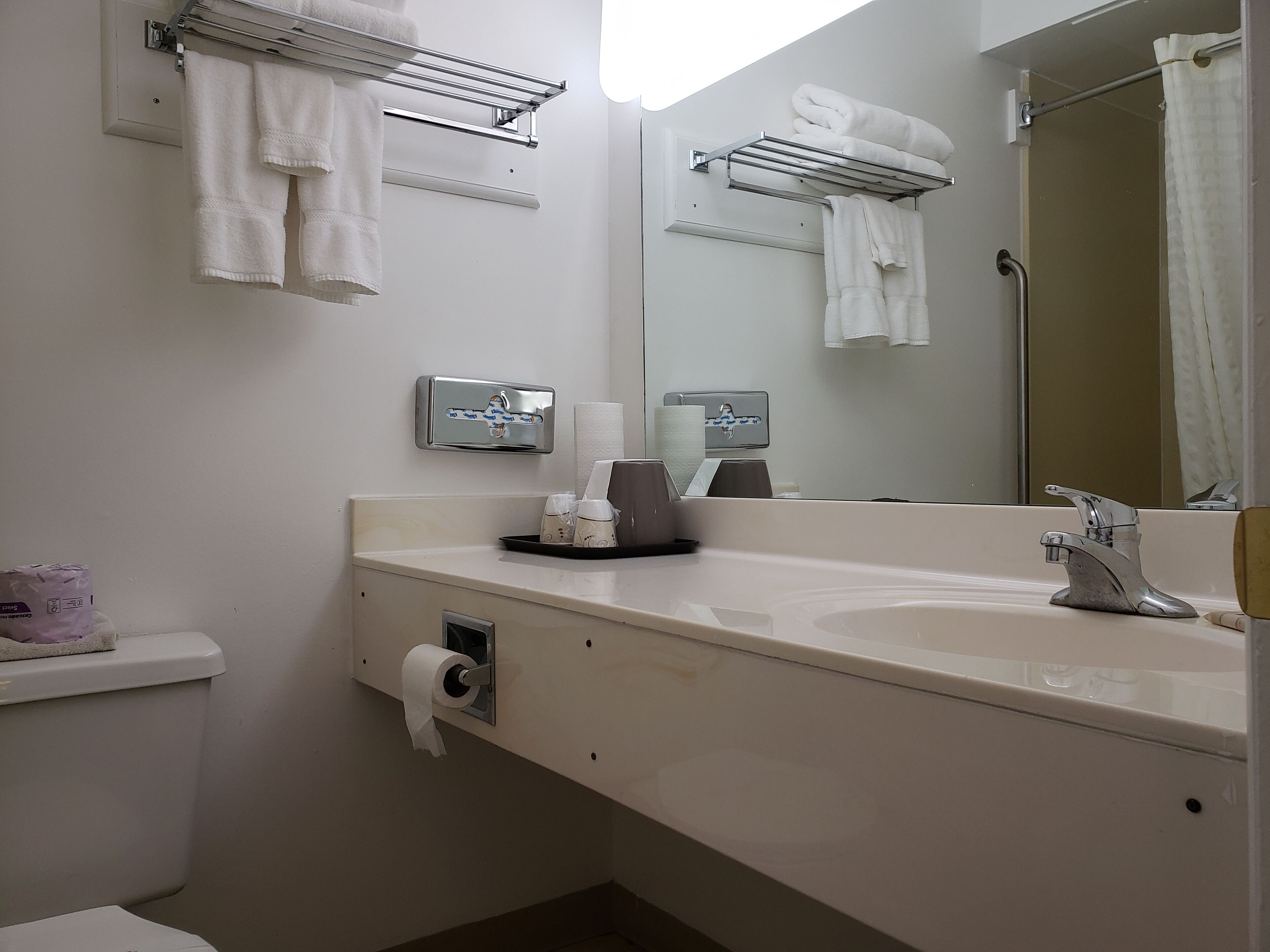 Un baño moderno con un lavamanos blanco, un espejo y un inodoro. Hay toalleros sobre el lavamanos y se ve una cortina de ducha al fondo.