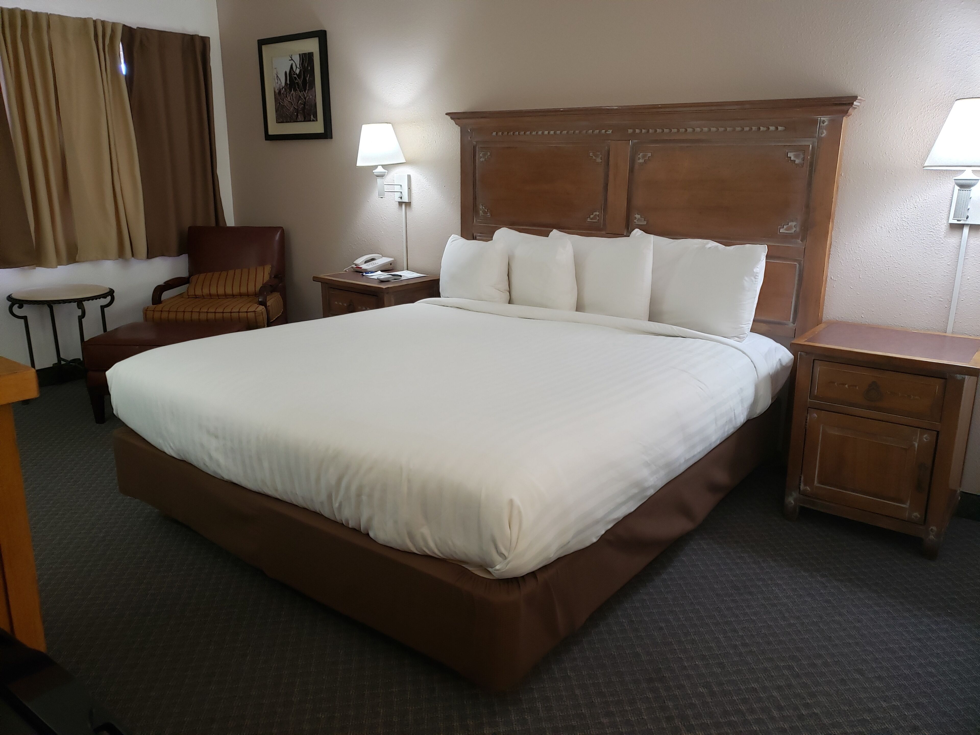 Une chambre d’hôtel avec un grand lit, une tête de lit en bois, une table de chevet avec une lampe, une chaise et un petit bureau.