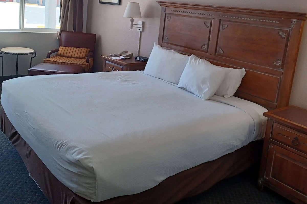 A large bed with white linens, a wooden headboard, a bedside table with a lamp, a chair, and a small round table.