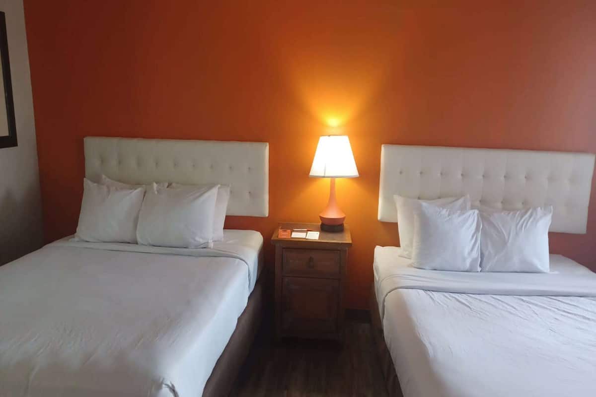 A hotel room with two beds, each with white linens and headboards, a nightstand with a lamp, and a wooden floor.