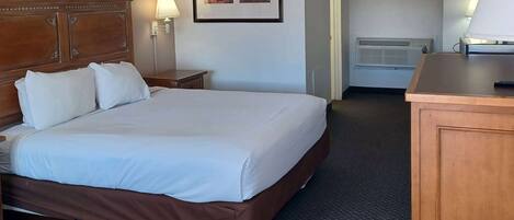 A hotel room with a large bed, a wooden headboard, a nightstand with a lamp, a framed picture on the wall, and a desk with a lamp.