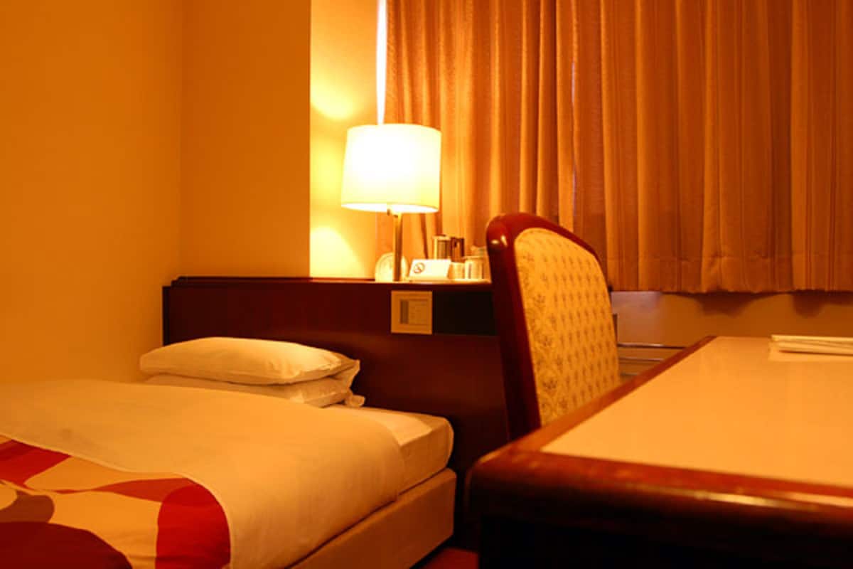 A hotel room with a bed, a chair, a nightstand with a lamp, and a window with curtains.