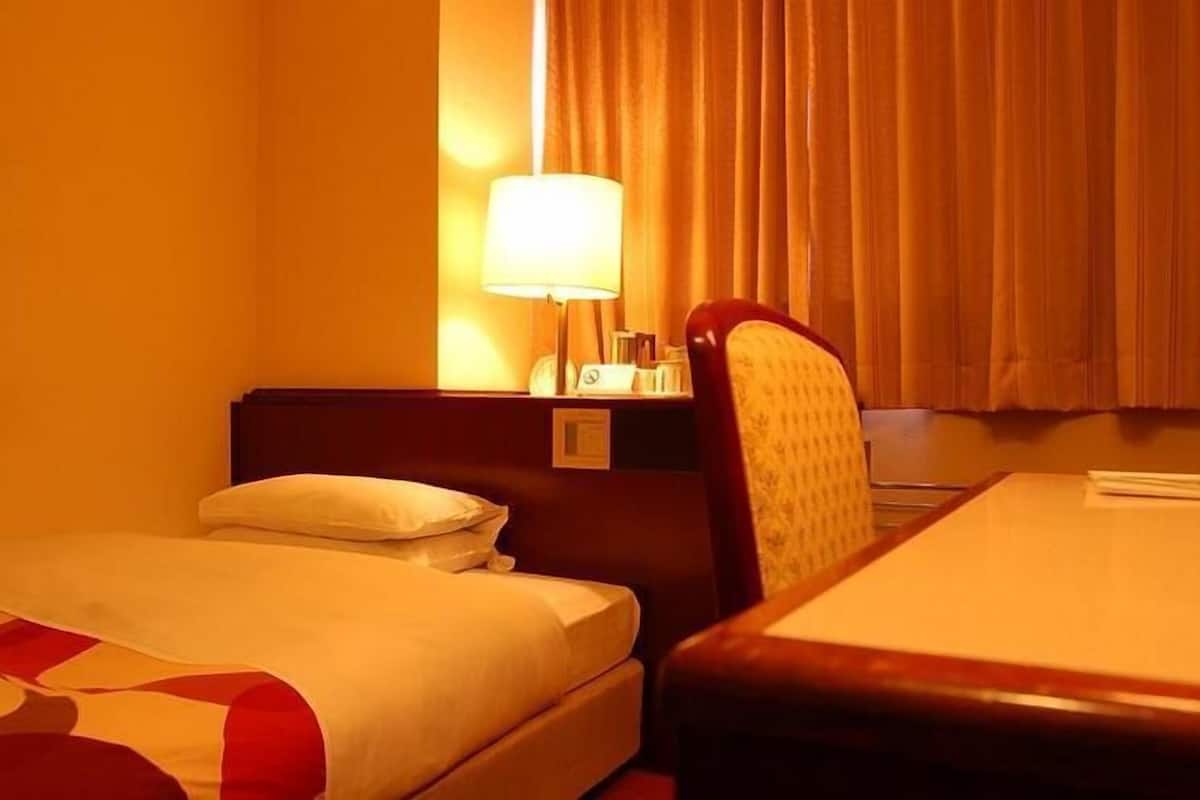 A hotel room with a bed, a desk, a chair, a lamp, and a window with curtains.