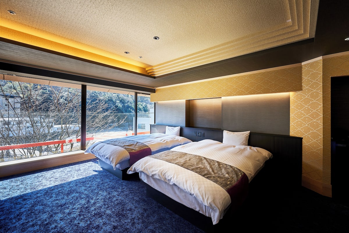 Een moderne hotelkamer met twee bedden, een groot raam met uitzicht op bomen, en een balkon met een rode leuning.