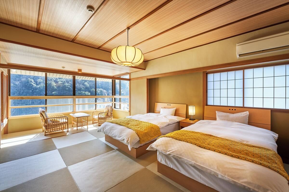 Hotelkamer met twee bedden, een zithoek met een tafel en stoelen, grote ramen met uitzicht en een traditioneel Japans plafond.