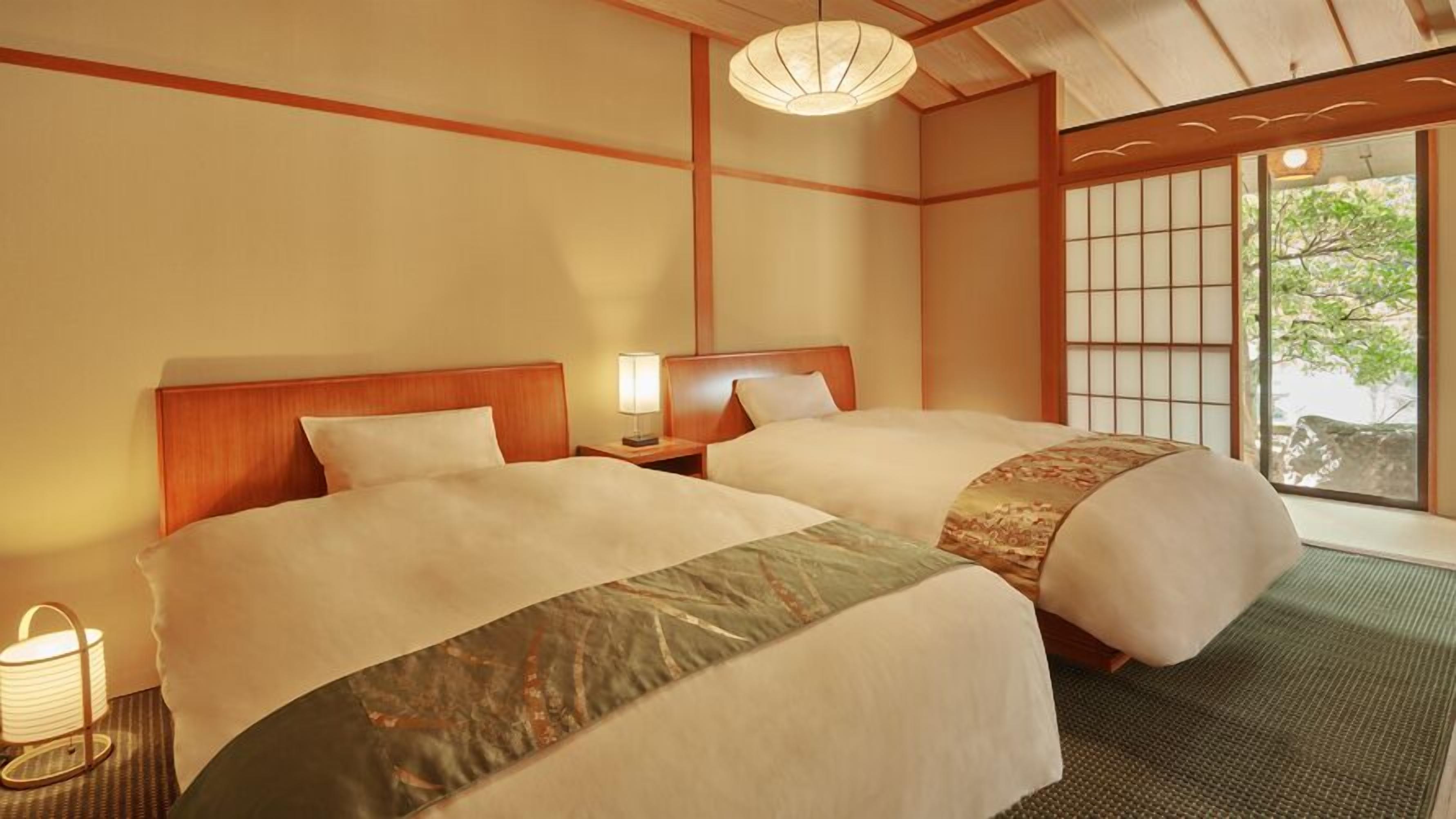 Une chambre d’hôtel de style japonais traditionnel, avec un sol recouvert de tatamis, deux lits dotés de têtes de lit assorties, un paravent shoji et une vue sur la verdure environnante.