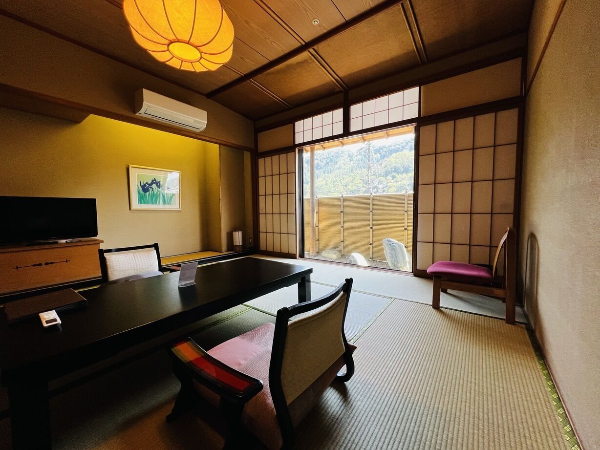 Une pièce de style japonais traditionnel, avec un sol recouvert de tatamis, une table basse, des chaises, un téléviseur et une vue sur l’extérieur.