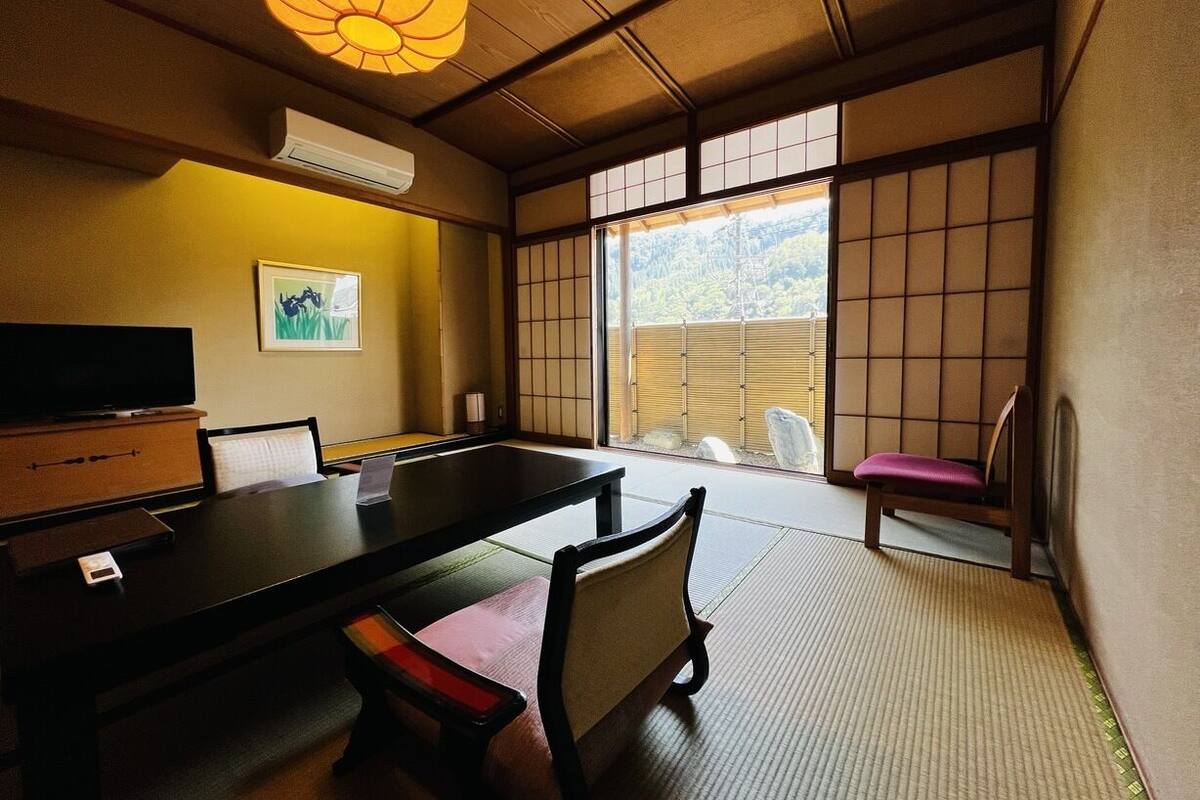 Een traditionele Japanse kamer met tatami vloerbedekking, een lage tafel, stoelen, een televisietoestel en uitzicht op het omliggende landschap.