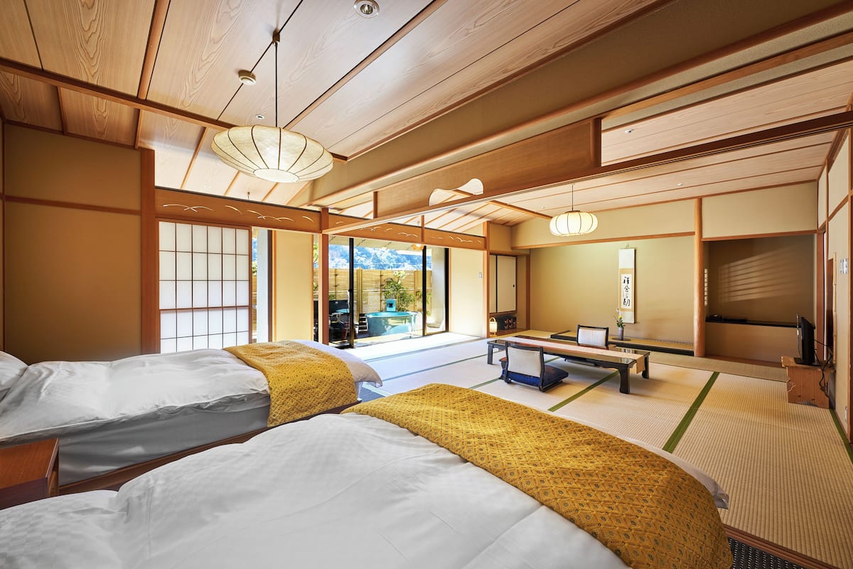 Een traditionele Japanse kamer met tatami vloer, twee bedden met wit en geel beddengoed, een houten plafond en uitzicht op het landschap door grote ramen.