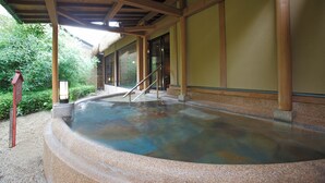 Ein traditionelles japanisches Onsen mit Holzdach, Steinbecken und einer Holztreppe, die zu einem überdachten Eingang führt.