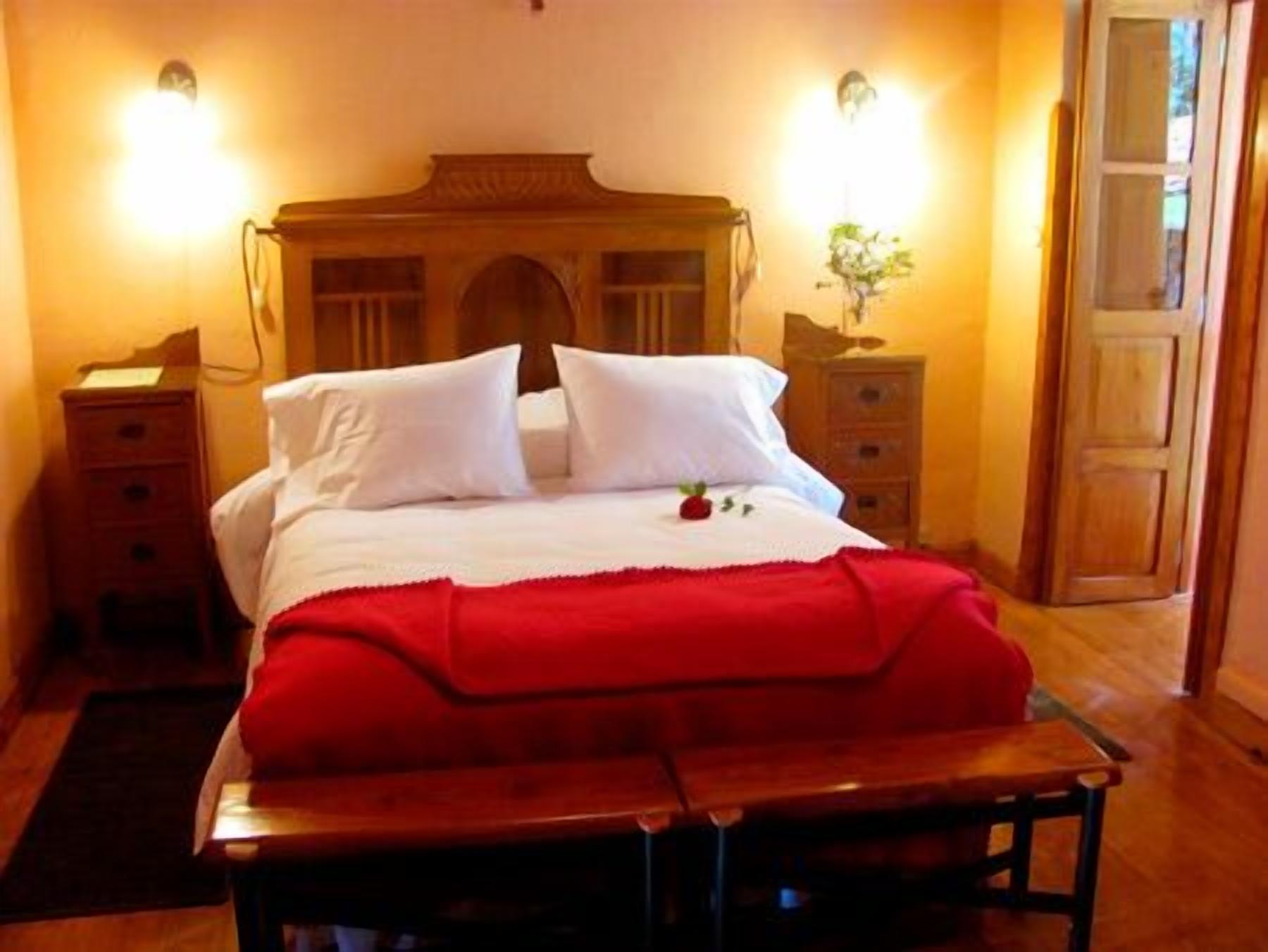 Una cama impecable con sábanas blancas y una colcha roja, flanqueada por mesitas de noche de madera con lámparas.