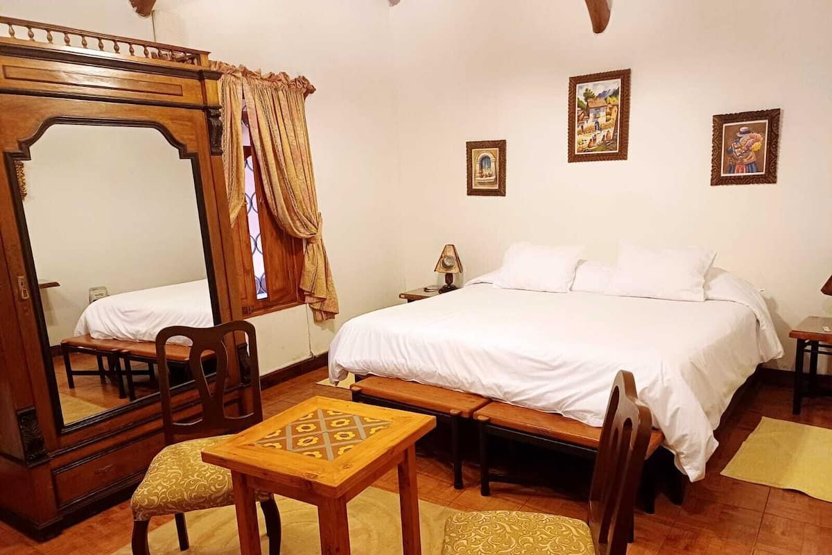 Un dormitorio con un espejo grande, una cama con sábanas blancas, una silla de madera, una mesita y una mesita de noche con lámpara.