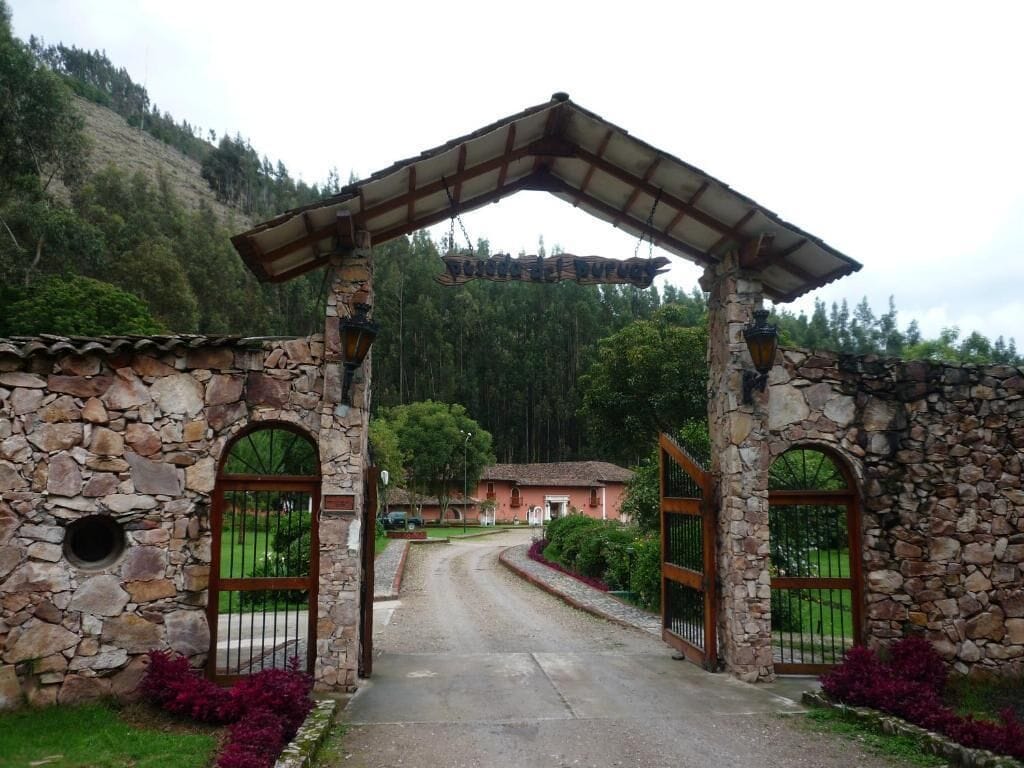 Un arco in pietra conduce a un edificio con tetto a tegole, immerso nel verde con montagne sullo sfondo.
