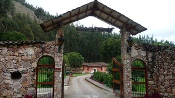 Un arco in pietra conduce a un edificio con tetto a tegole, immerso nel verde con montagne sullo sfondo.