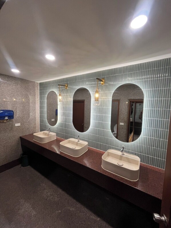 A public restroom with three sinks, each with a mirror above. The walls are tiled with a vertical pattern, and there are recessed ceiling lights.