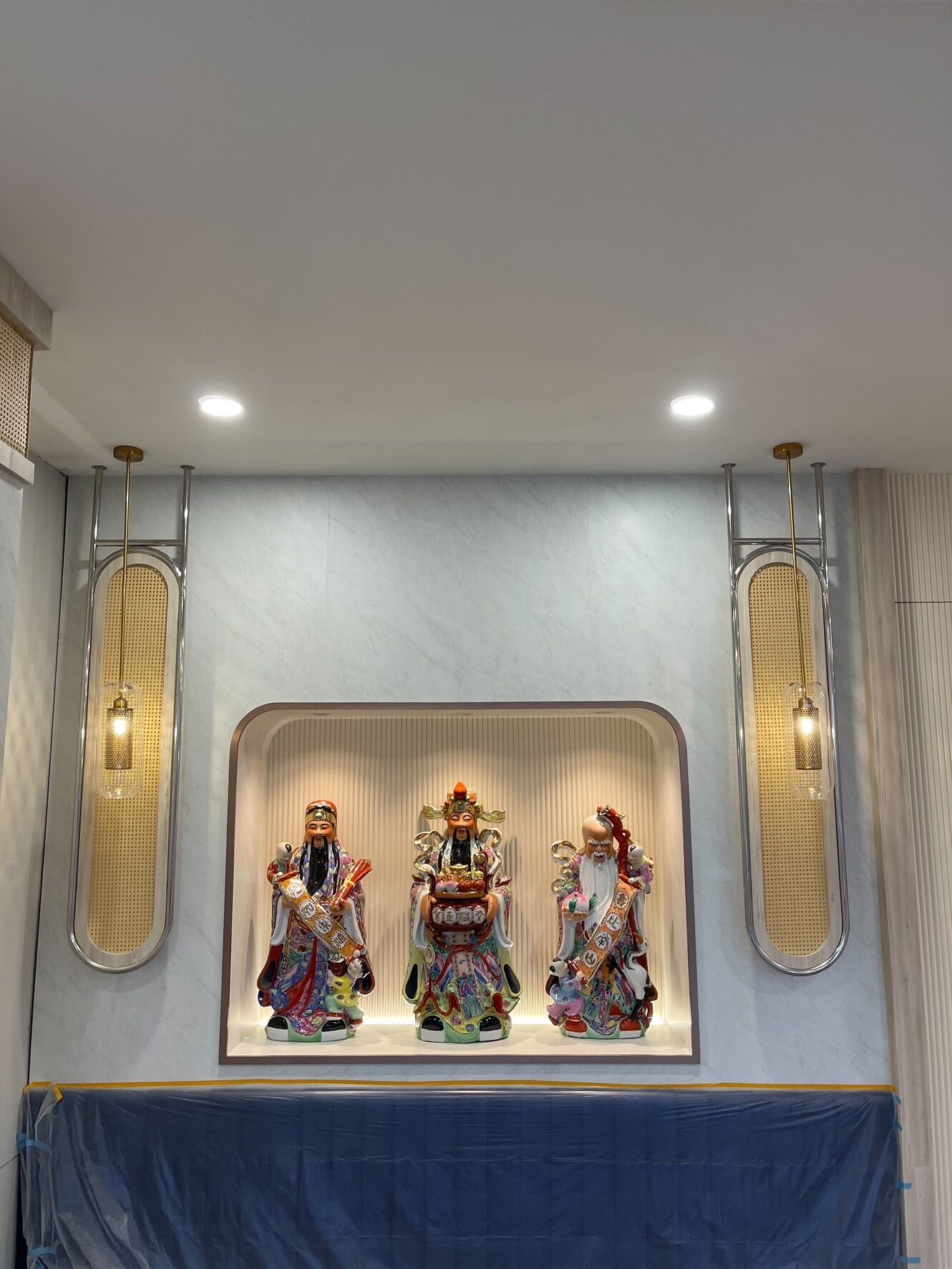 A wall-mounted display of three ornate statues in traditional attire, with two wall-mounted lights on either side.