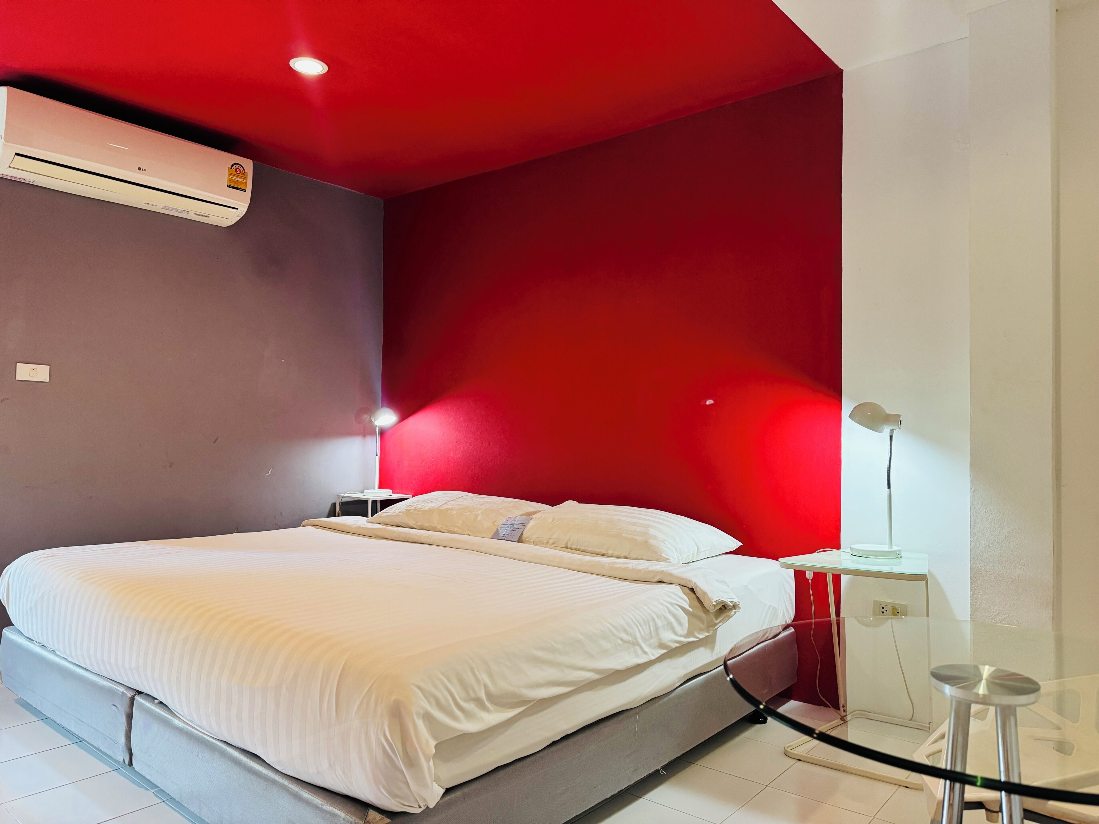 A bedroom with a bed, an air conditioner, a nightstand, a lamp, and a small table.