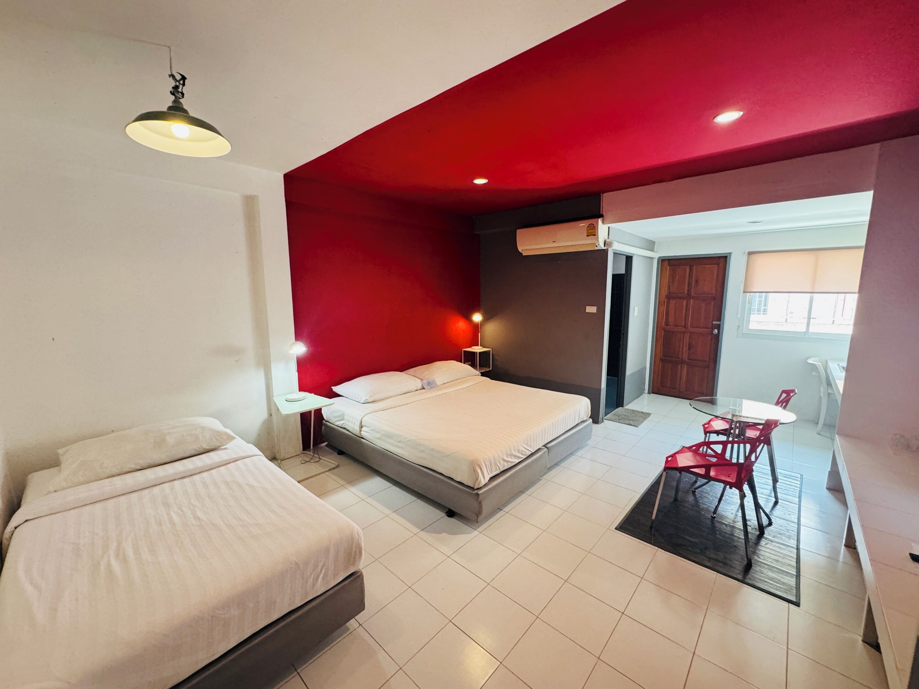 Habitación de hotel con dos camas, una pared de acento roja, una zona de comedor con sillas rojas y una ventana.