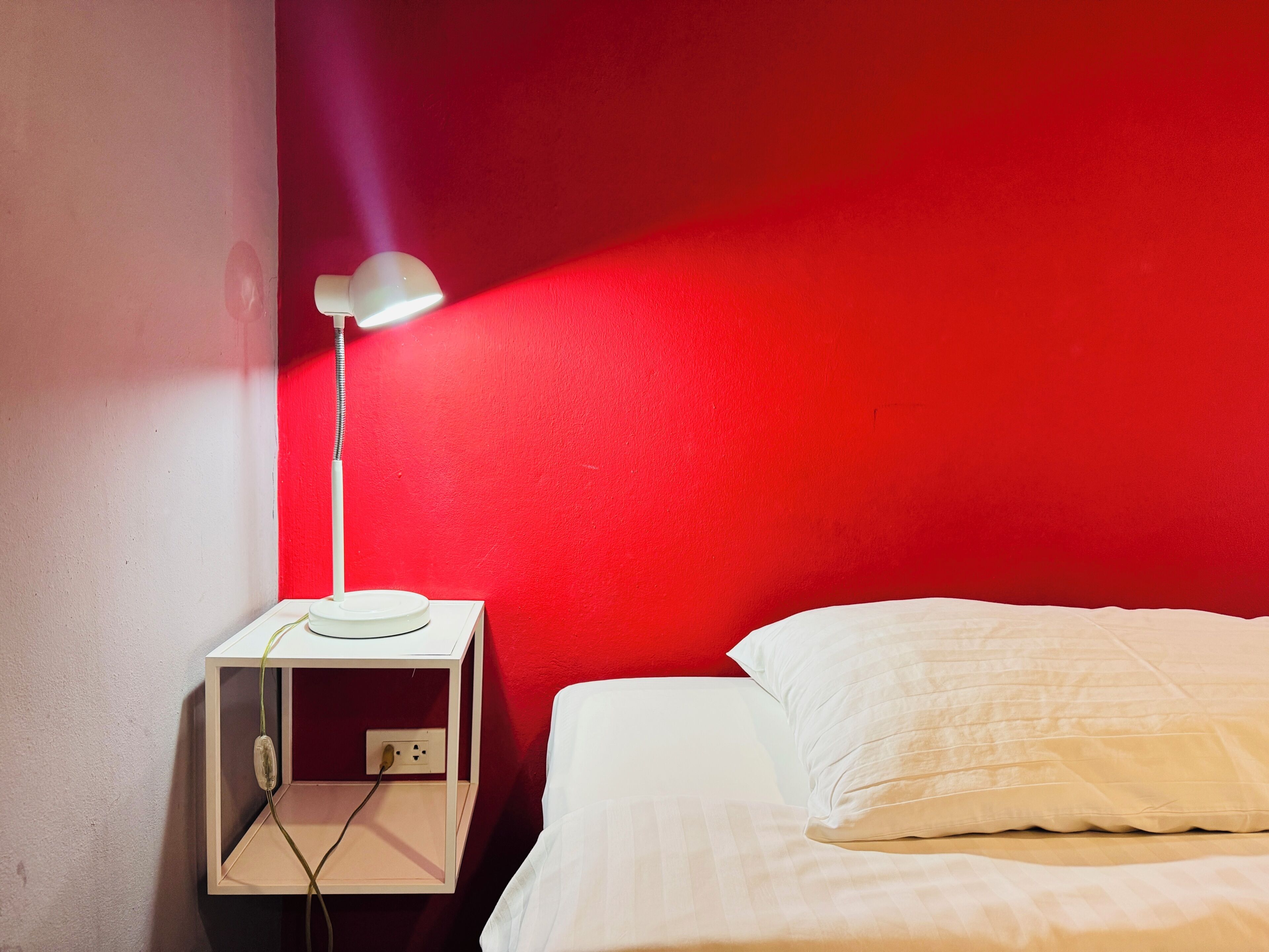 A bed with white bedding, a white bedside table with a lamp, and a red wall.