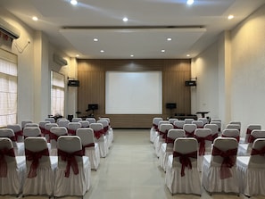 Un salón de conferencias con filas de sillas, una pantalla de proyector y luces en el techo.