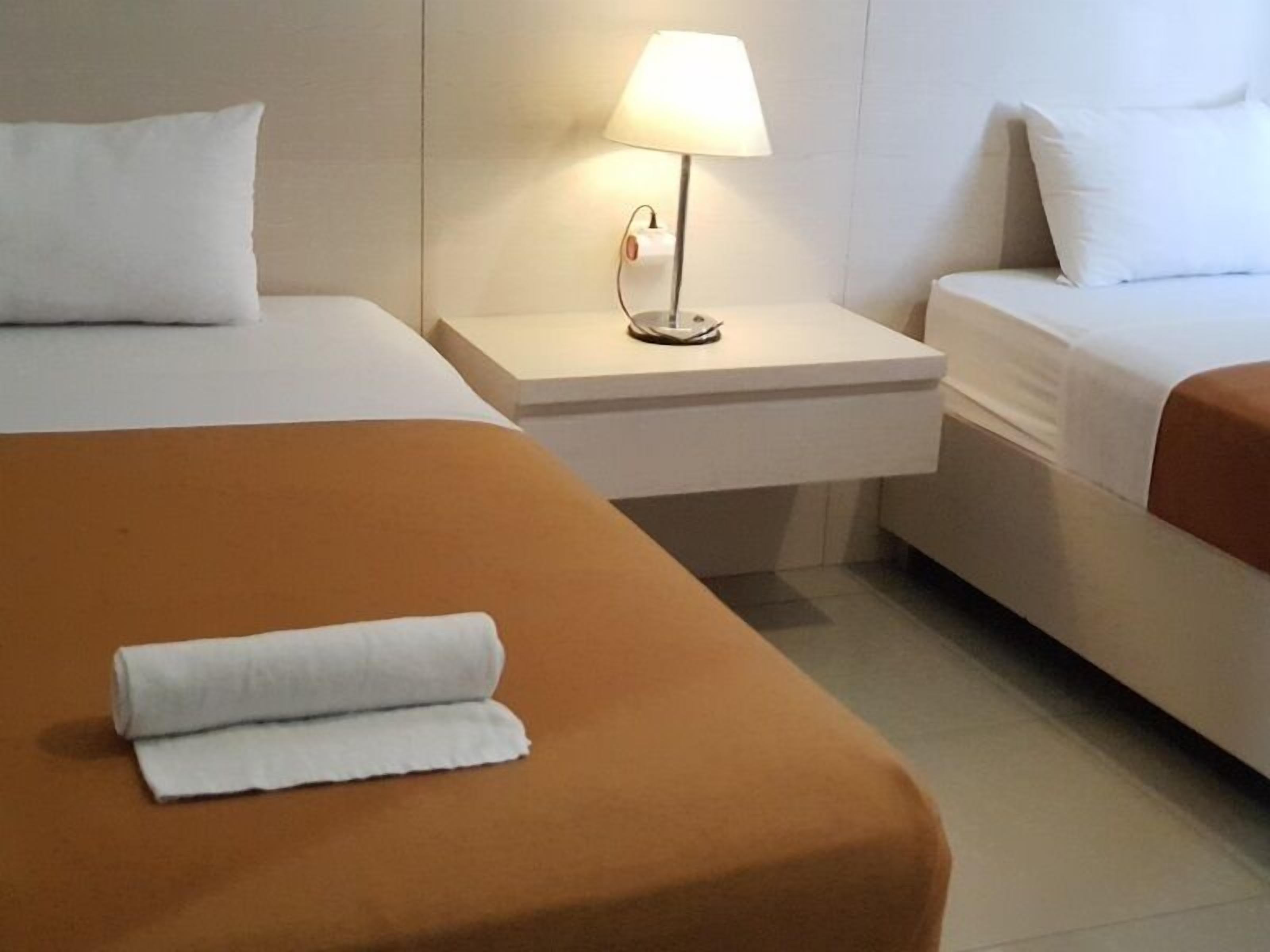 A hotel room with two beds, one with a brown bedspread and the other with a white bedspread. There is a nightstand with a lamp between the beds.