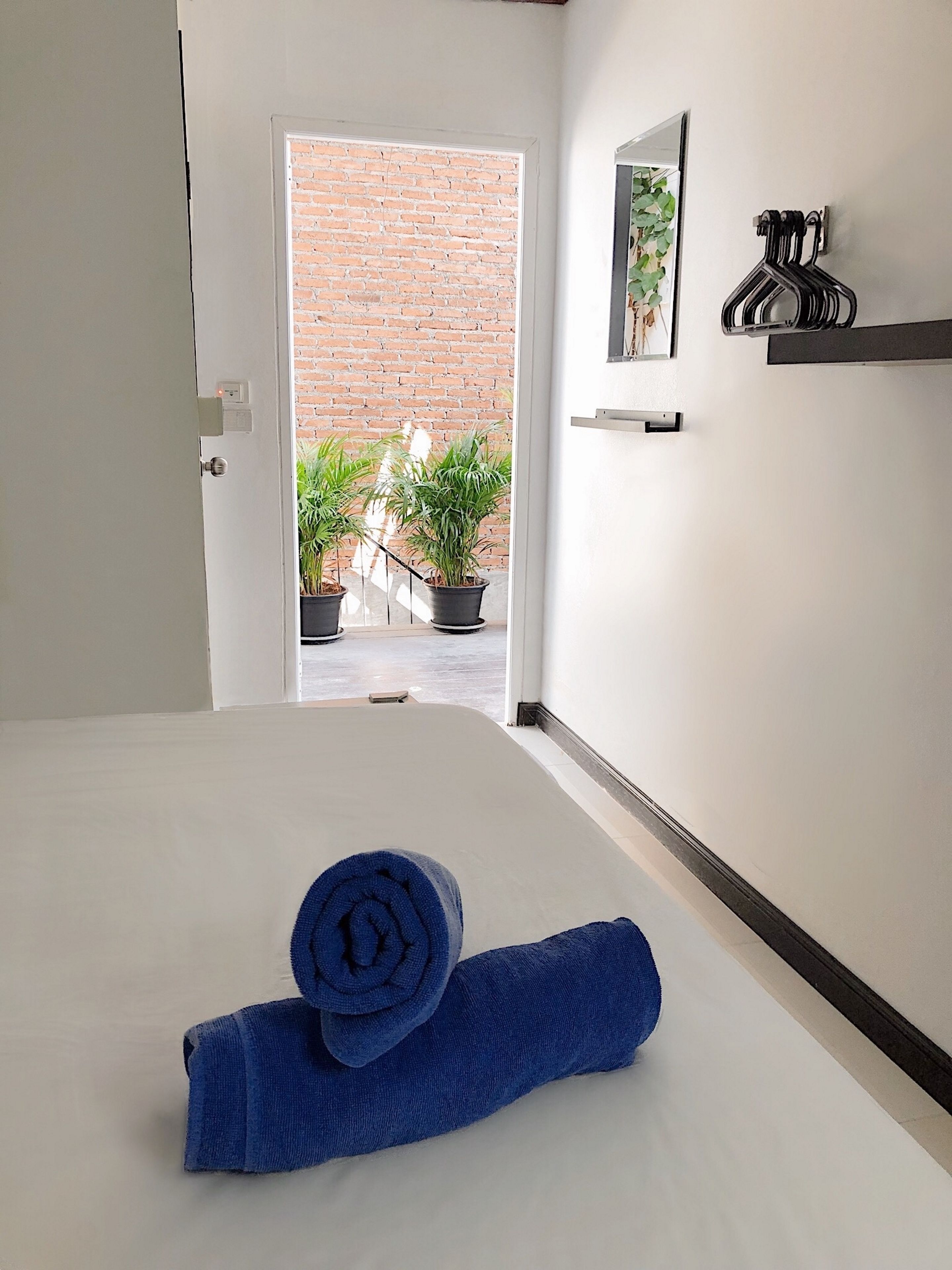 Habitación de hotel con una cama, una puerta que da a un muro de ladrillo, plantas en macetas y un estante fijado a la pared con una toalla y un cuadro.