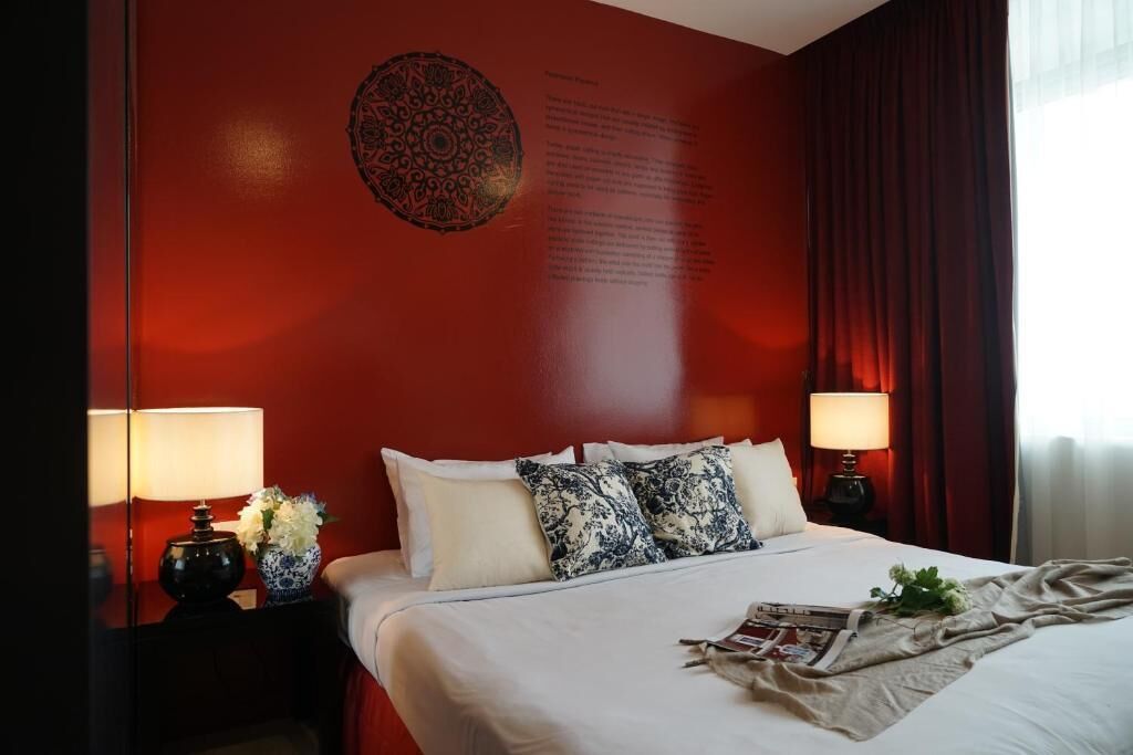 A hotel room with a red accent wall, a large bed with white linens, two bedside tables with lamps, and a decorative wall hanging.