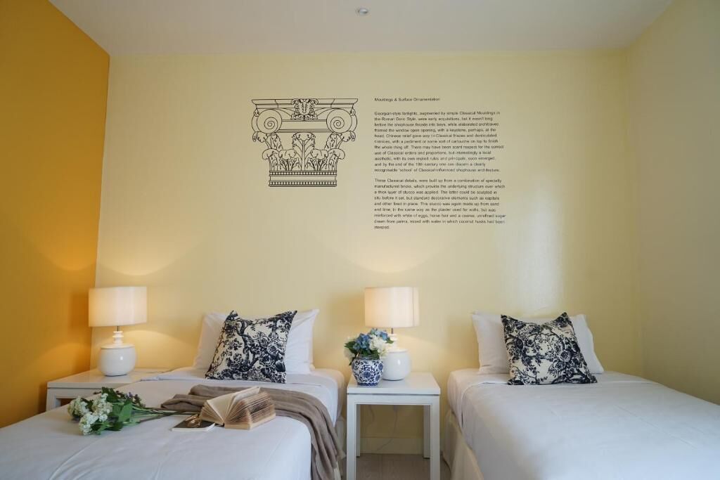 A hotel room with two beds, each with a bedside table, lamps, and decorative pillows. There is a wall-mounted informational plaque above the beds.