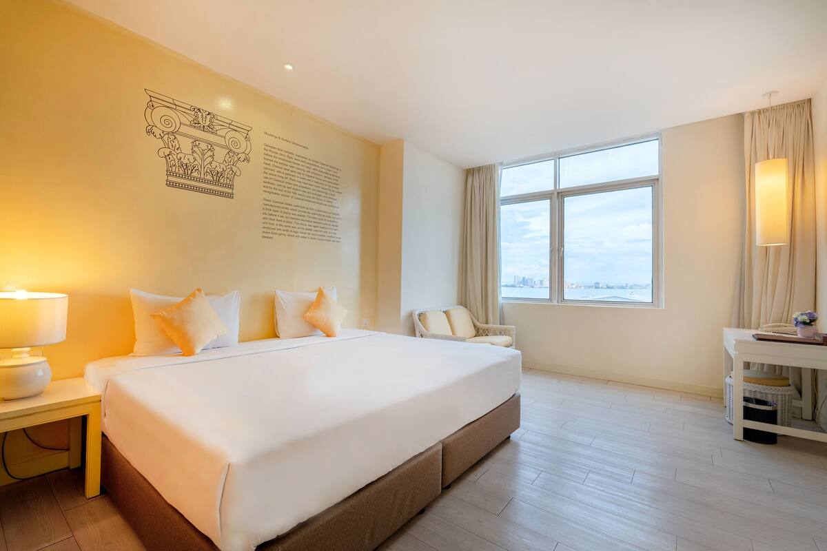 Habitación de hotel con una cama grande, un escritorio con lámpara, una silla y vistas a la ciudad por la ventana.