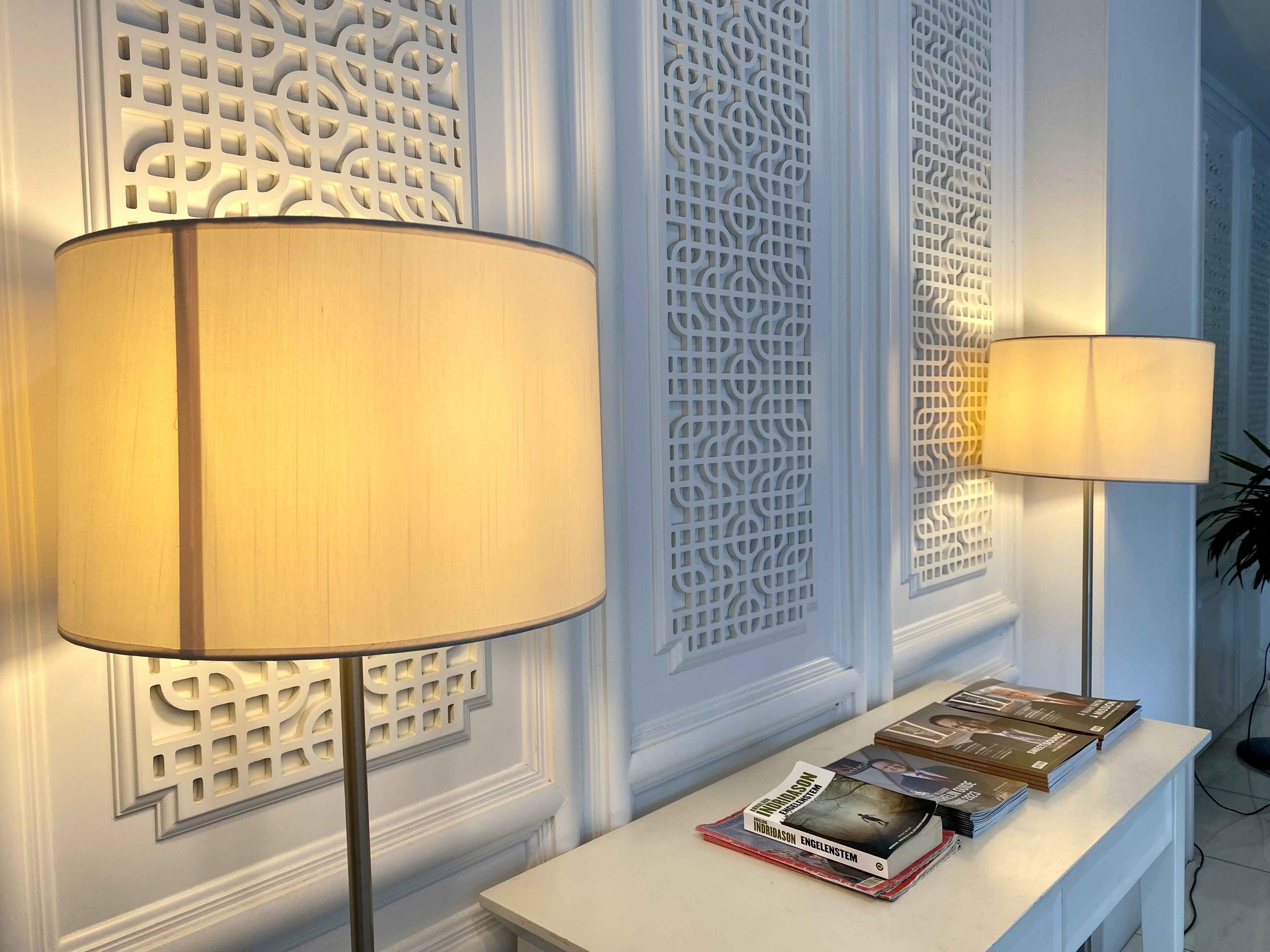 Two floor lamps with lampshades on a white console table, a decorative wall panel, and a stack of books.
