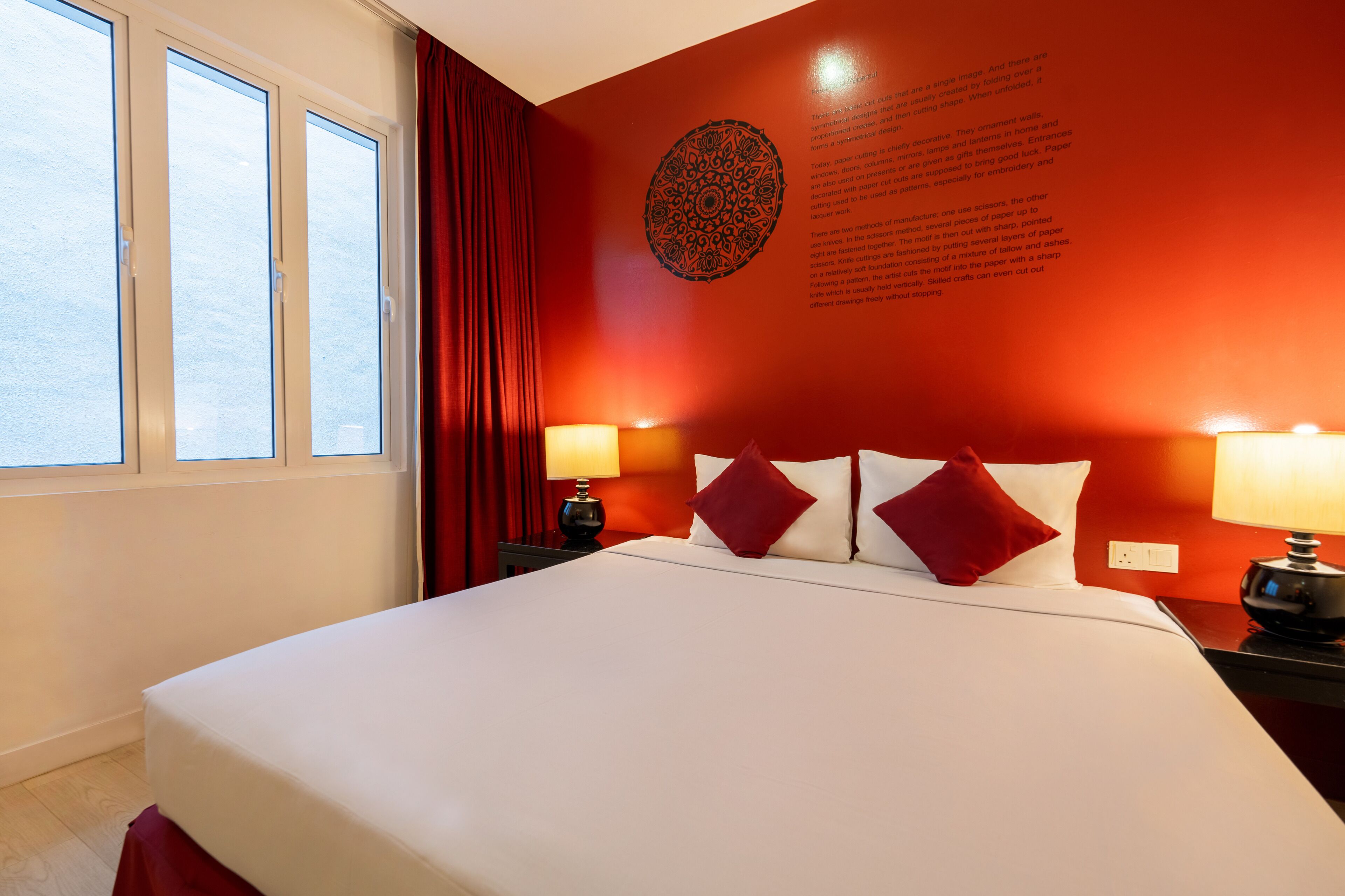 A hotel room with a large bed, red curtains, two bedside lamps, and a wall with a circular design and text.