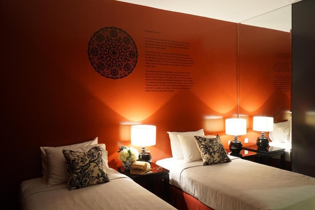 A hotel room with two beds, each with a nightstand, lamps, and decorative pillows. A large circular wall hanging is visible on the wall.