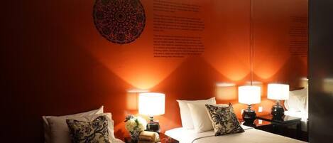 Habitación de hotel con dos camas, cada una con una mesita de noche, lámparas y almohadones decorativos. En la pared se ve un tapiz circular grande.