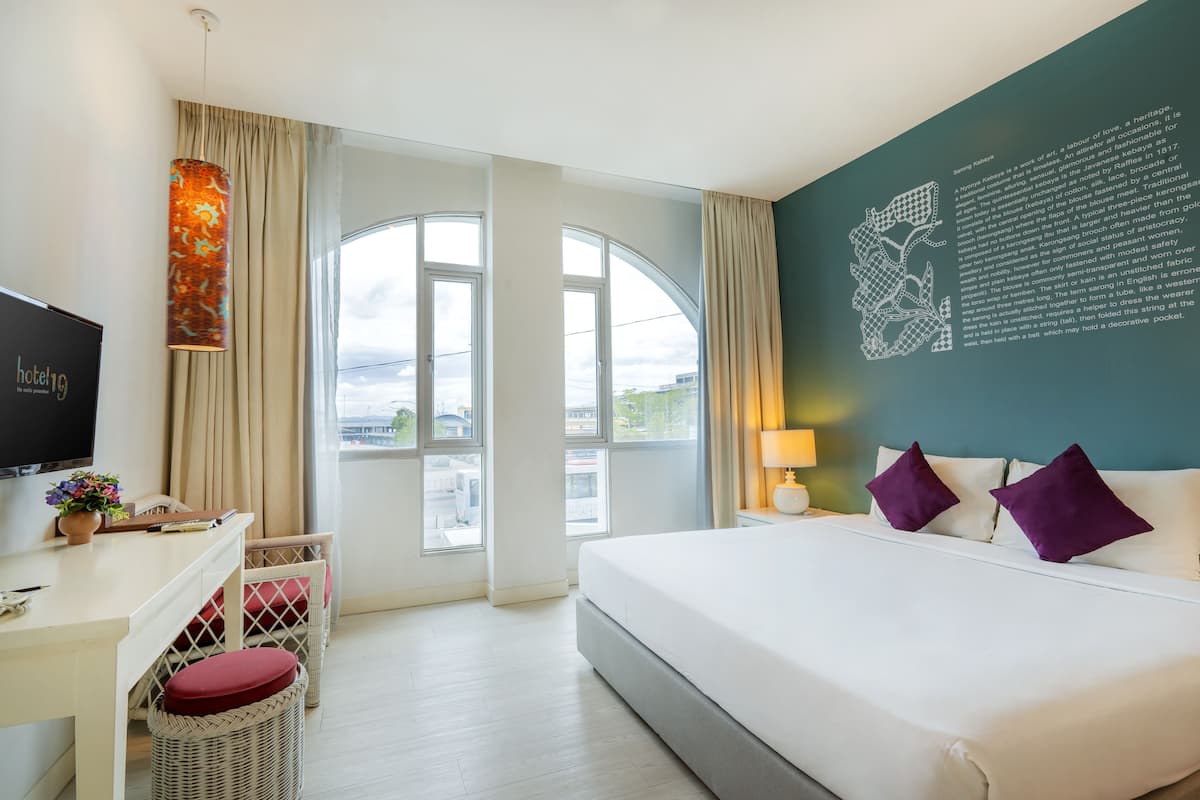 Habitación de hotel con una cama grande, un escritorio con una silla, un televisor y vistas de la ciudad a través de amplias ventanas.