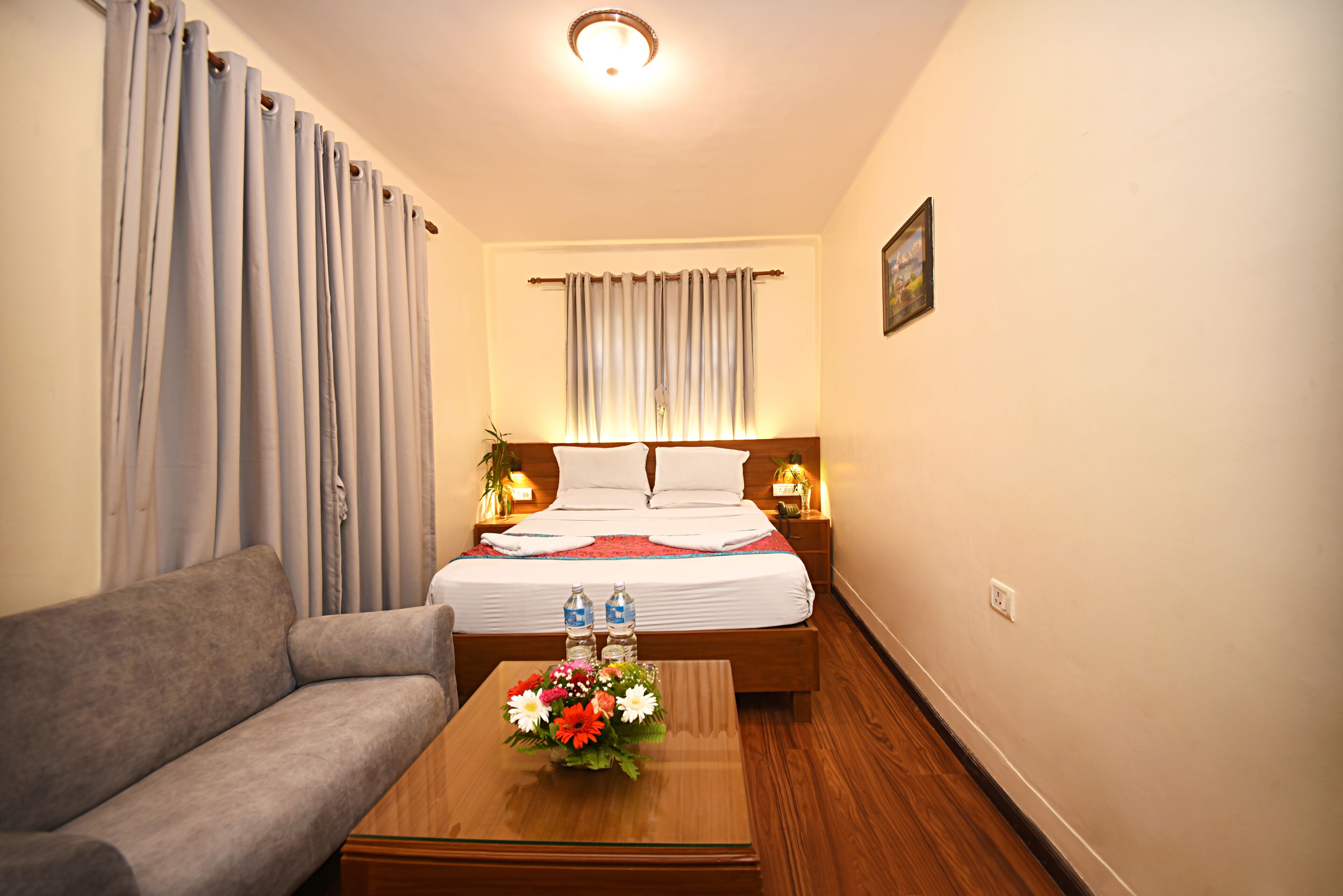A hotel room with a bed, a sofa, a wooden coffee table with flowers, a nightstand with a lamp, and a window with curtains.