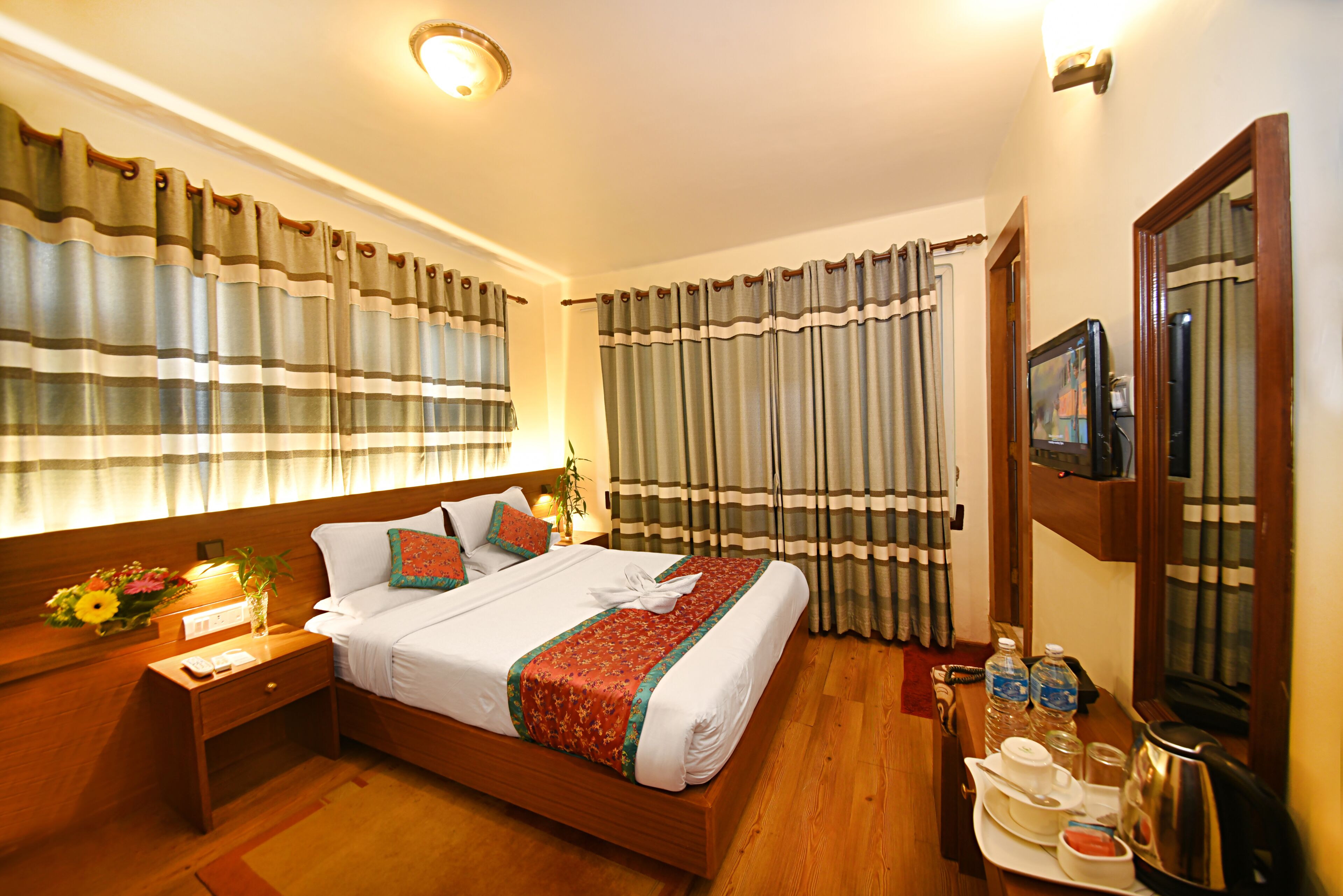 Habitación de hotel con cama de madera, cortinas, mesita de noche con lámpara, bandeja con alimentos para el desayuno y televisión en la pared.