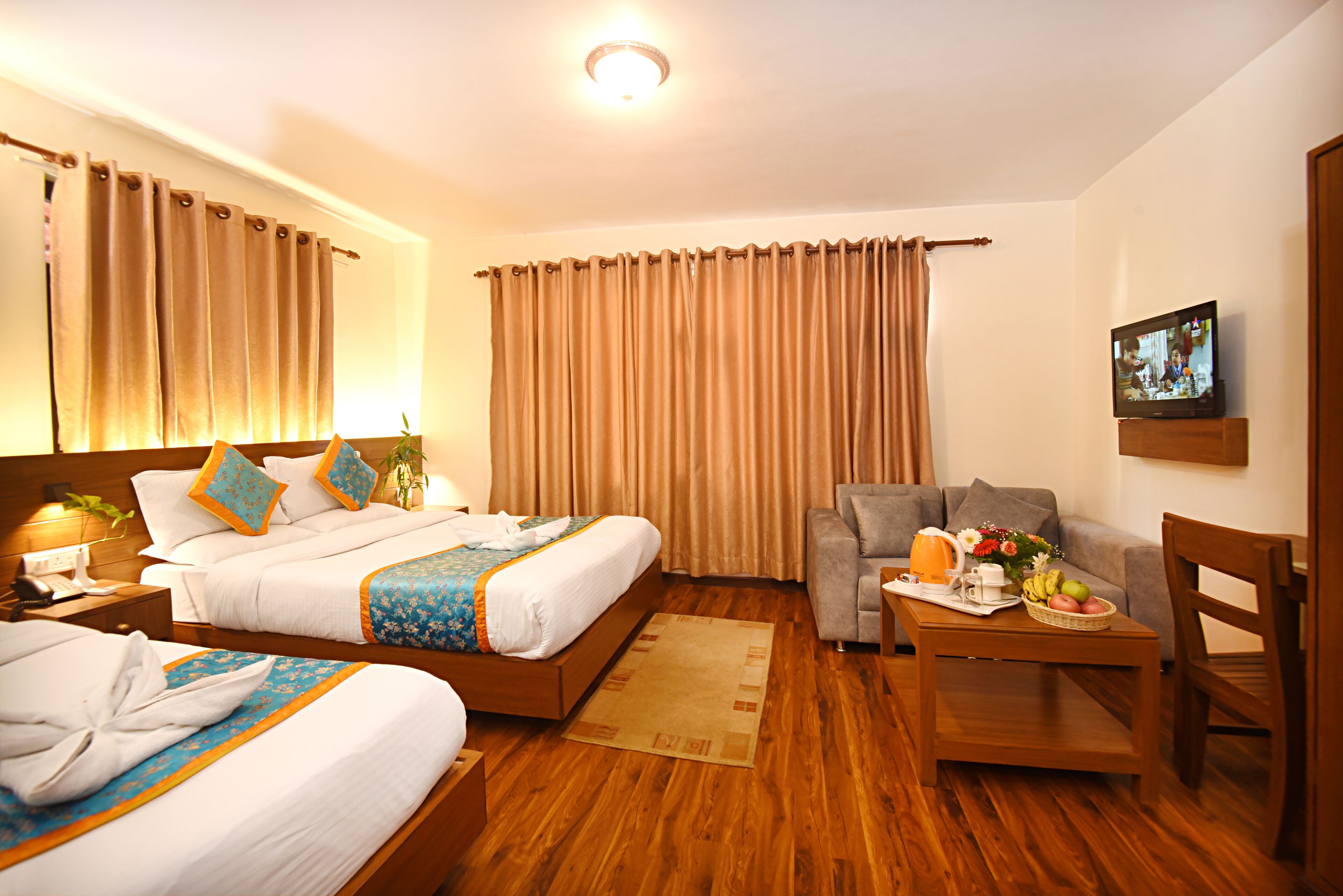 Quarto de hotel com duas camas, um sofá, uma pequena mesa com frutas e uma TV na parede.