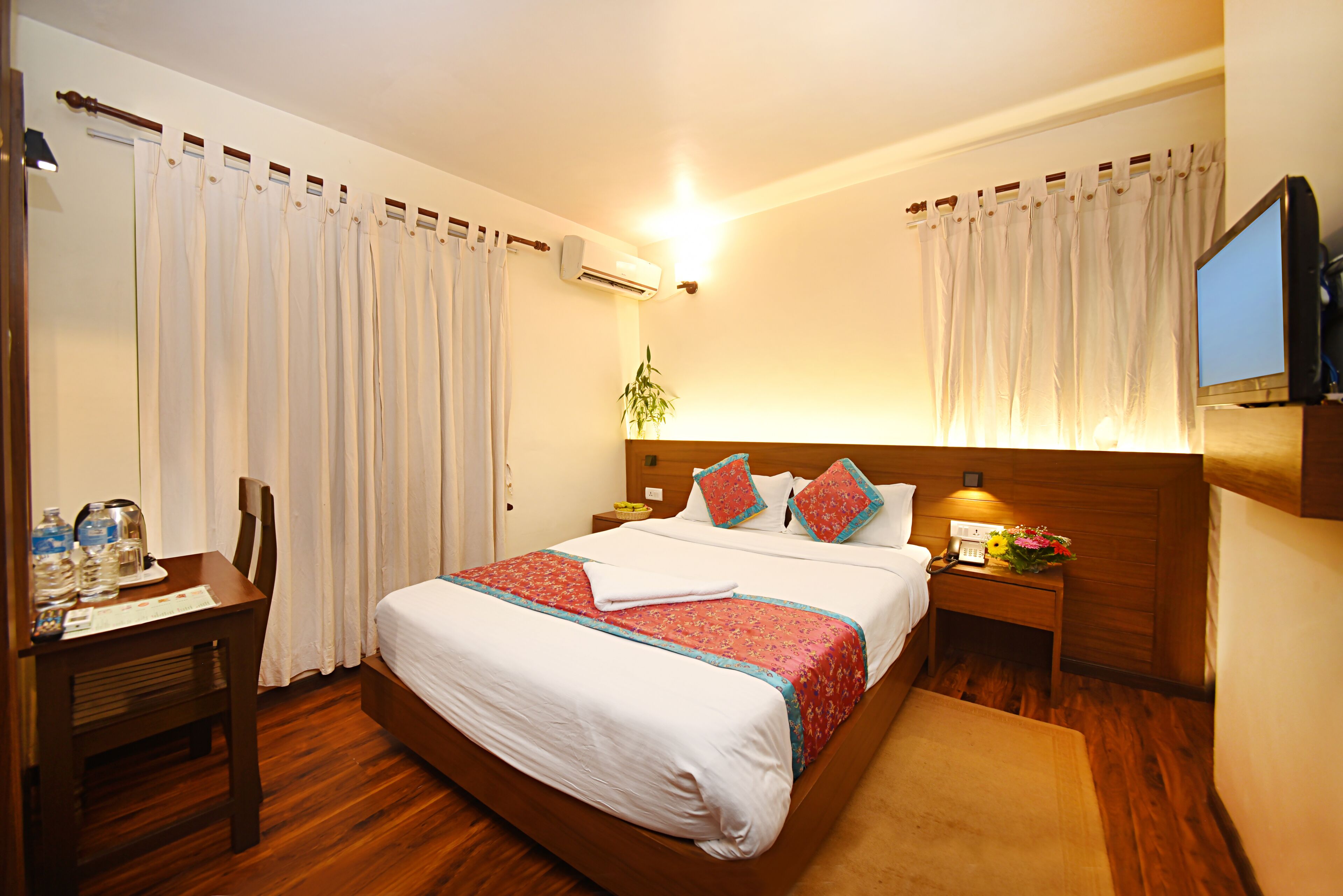 A hotel room with a wooden bed, white curtains, a desk with a chair, a bedside table with a lamp, and an air conditioner.