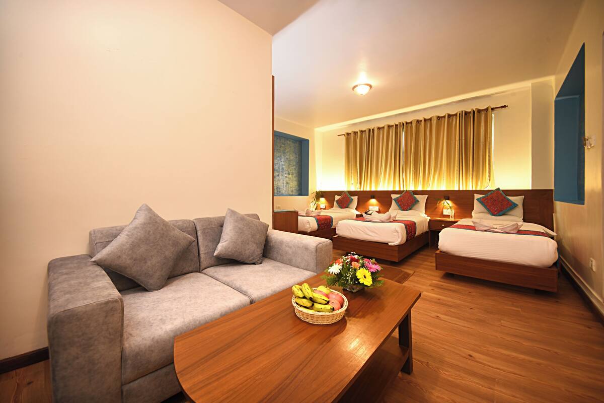 A hotel room with a grey sofa, a wooden coffee table with fruit and flowers, and two beds with headboards and pillows.