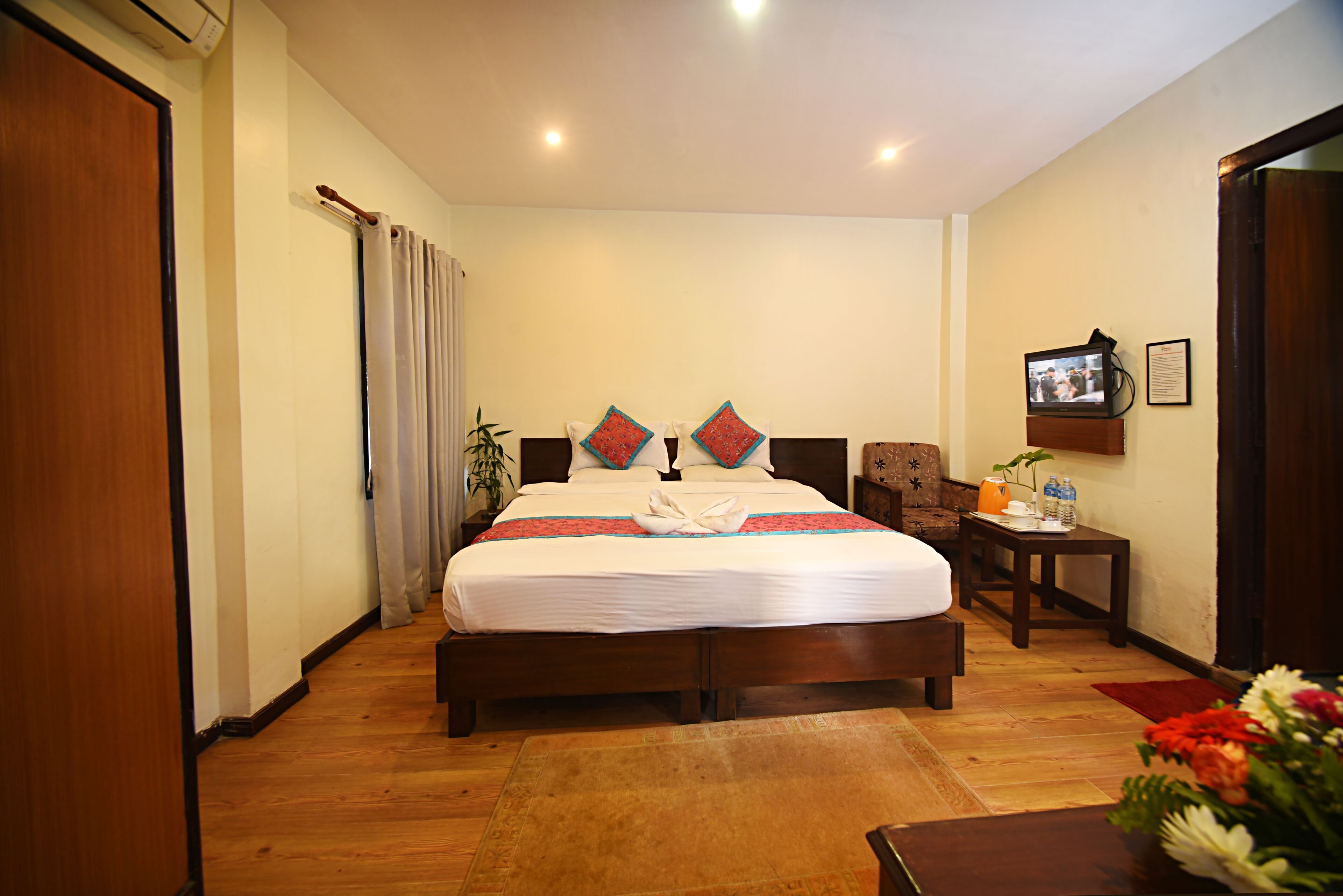 Una habitación de hotel con una cama grande, suelo de madera, una pequeña mesa con una planta y un televisor en la pared.