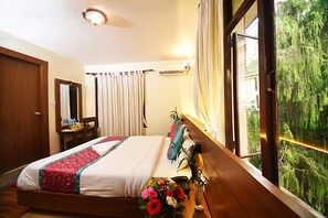 A hotel room with a bed, a bedside table with a vase of flowers, a window with curtains, and a view of trees outside. - Hotel Amaryllis (Kathmandu)