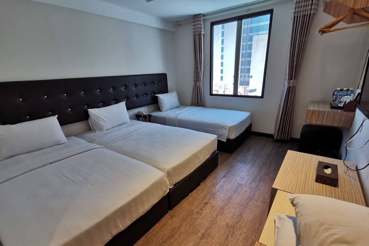 A hotel room with two beds, a wooden floor, a large window with curtains, and a desk with a telephone.