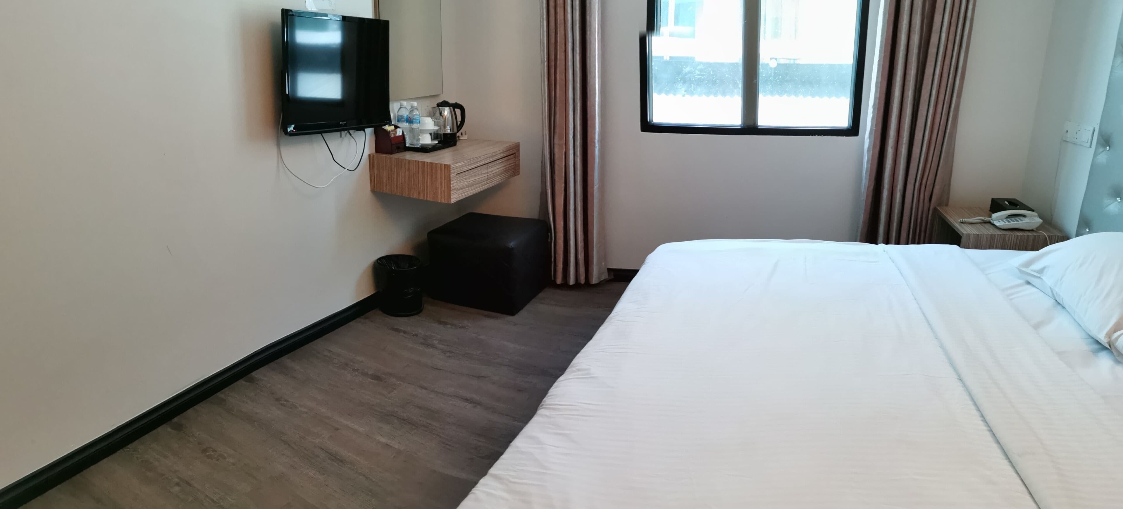 A hotel room with a bed, a TV mounted on the wall, a small wooden side table, a trash bin, and a window with curtains.