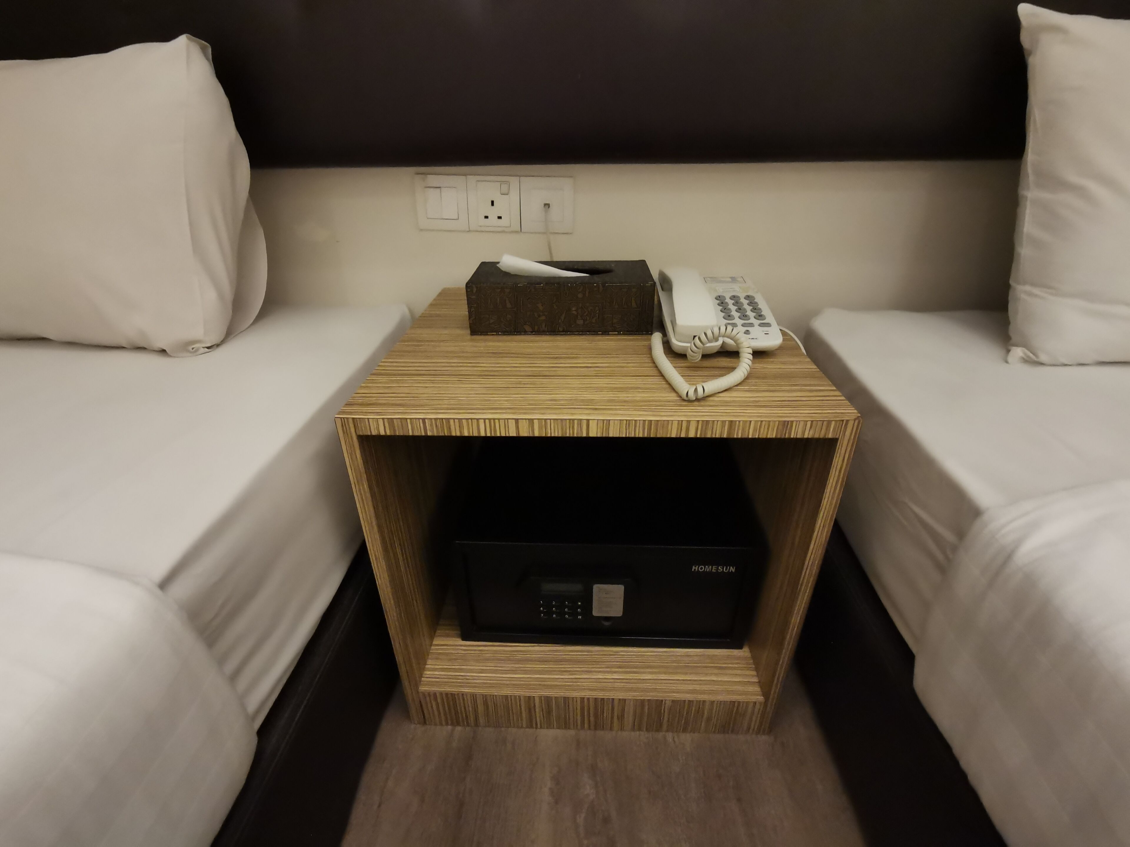 Habitación de hotel con dos camas, una mesita de noche de madera con un teléfono y una caja, y un dispositivo electrónico negro sobre la mesita.