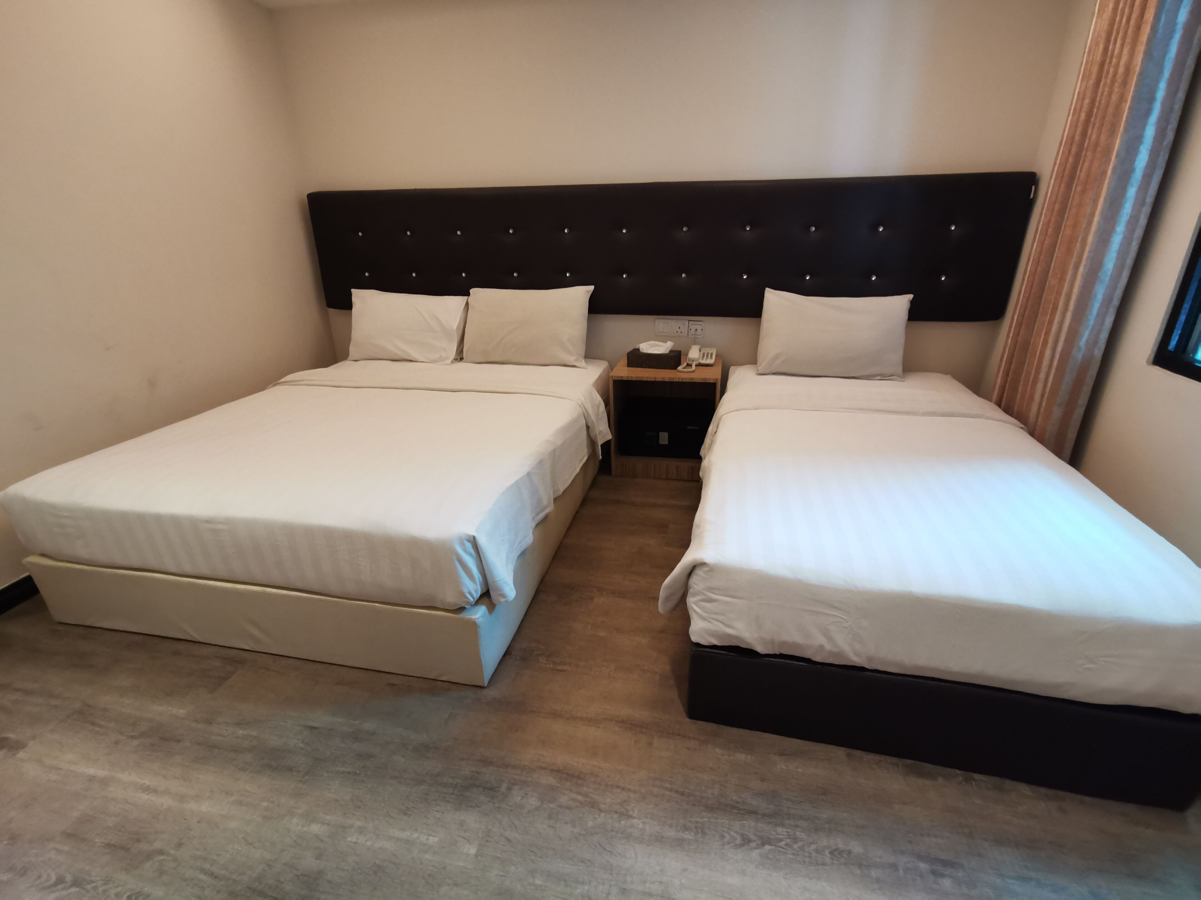 Quarto de hotel com duas camas de solteiro, cabeceira escura, mesa de cabeceira com telefone e lenços, e piso de madeira.
