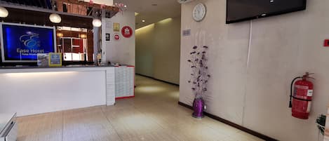 Zona de recepción de hotel con mostrador, televisor montado, reloj de pared y extintor.