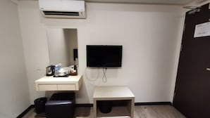 A hotel room with a white air conditioner, a flat-screen TV mounted on the wall, a small wooden cabinet, a black leather stool, and a white sink with toiletries. - Ease Hotel (Kota Kinabalu)