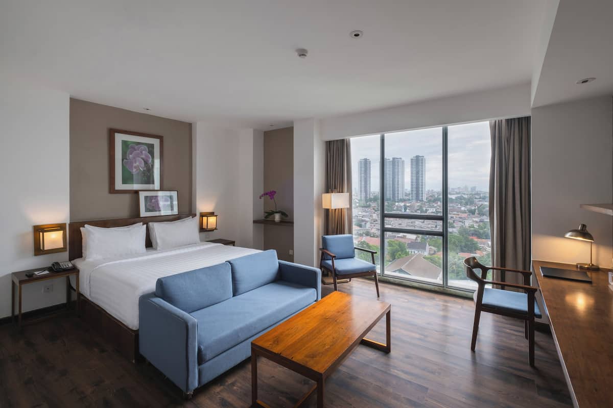A hotel room with a large bed, a blue sofa, a wooden coffee table, and a desk. There are two chairs, a window with a city view, and a small plant on a shelf.